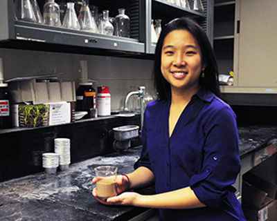 A photo of a young woman in a lab holding a container of light brown dust