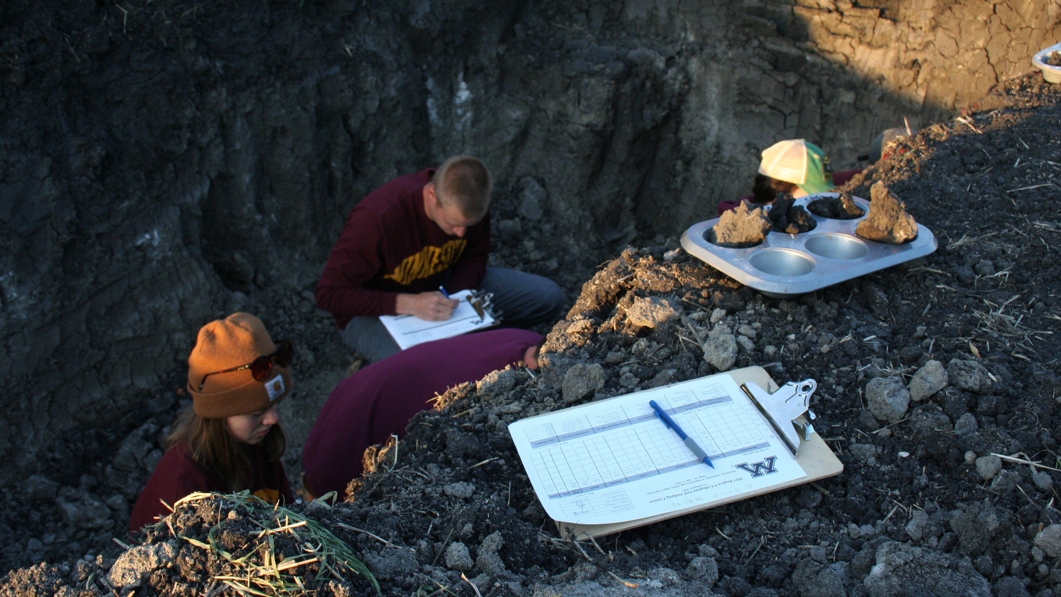 A photo looking down into a soil pit; a clipboard and a tin of soil samples are in the foreground; students in the pit are in the background. Photo by Aimee Gillespie.