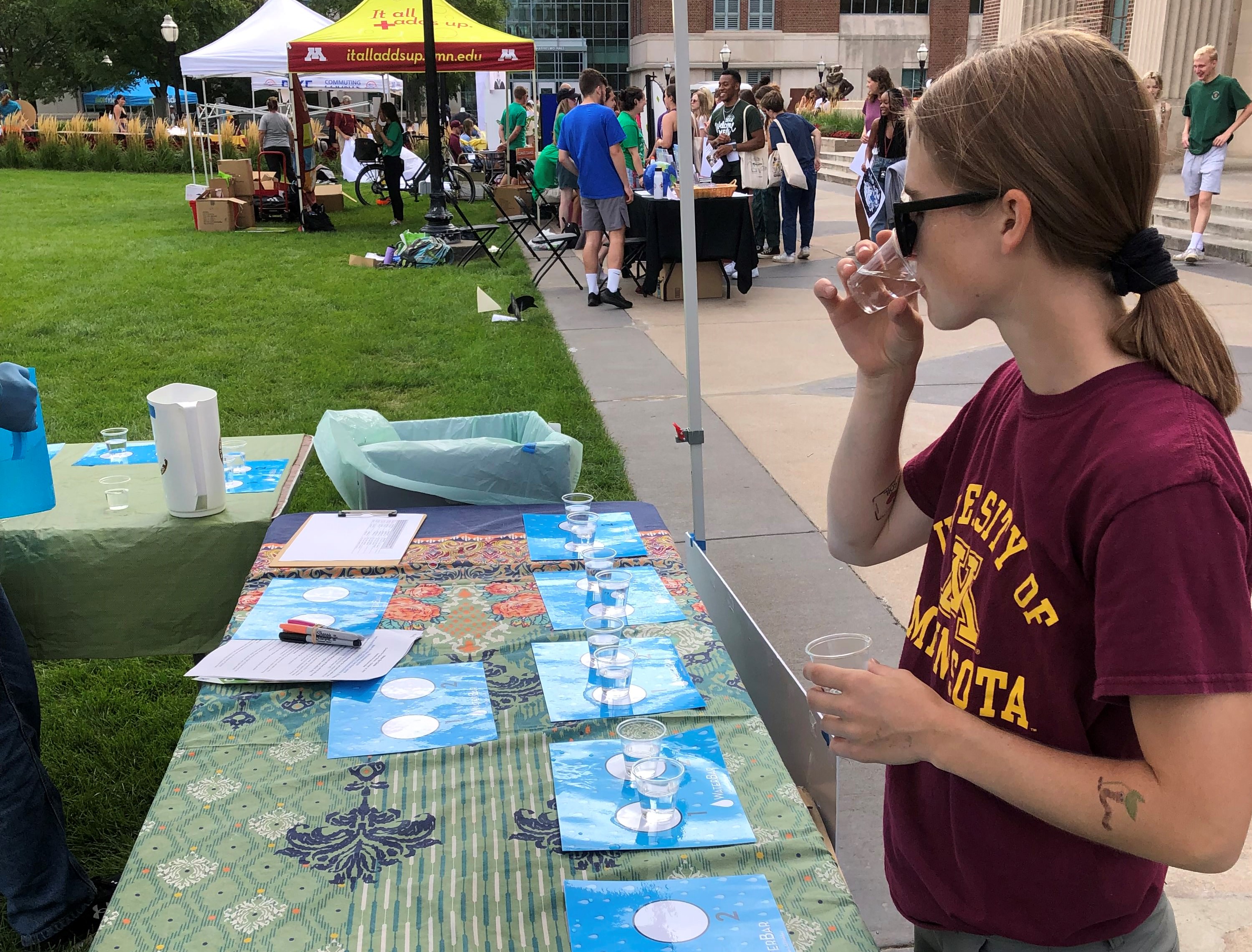 A photo of graduate student Bailey Tangen sipping two glasses of water at a student event called "The Water Bar"