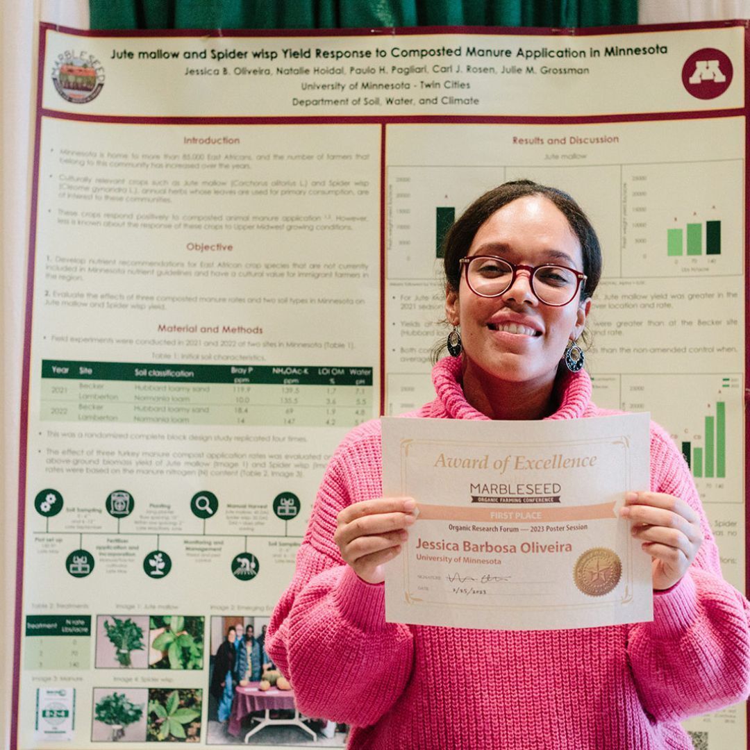A photo of Jessica Barbosa Oliveira holding her award in front of her research poster