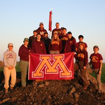 A photo of the 2021 Soil Judging Team in Crookston, Minnesota standing on a pile of soil holding a University of Minnesota flag