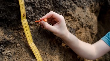 A close up photo of a soil pit with a tape measure and a hand placing a marker at a color change horizon; photo by Liz Banfield