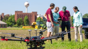 A photo of a drone in the foreground, four researchers standing in a field in the midground, and the St. Paul campus water tower in the background; photo by Krista Styer.