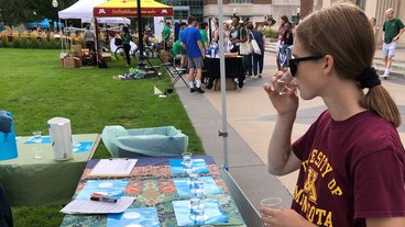 A photo of graduate student Bailey Tangen sipping two glasses of water at a table event called The Water Bar.