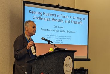 Carl Rosen, a man, stands in front of a podium and screen. The text on the screen reads: Keeping Nutrients in Place: a Journey of Challenges, Benefits, and Tradeoffs